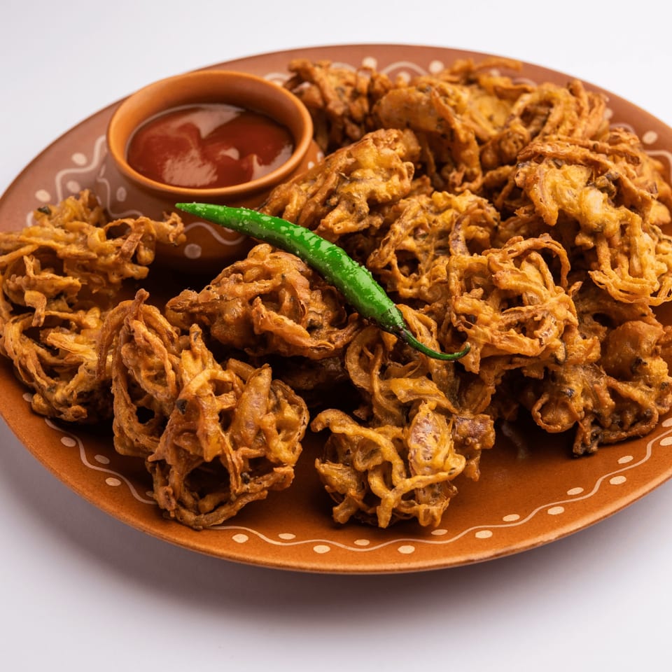 Crispy golden onion pakoras with a side of red chutney and a green chili on a brown plate.