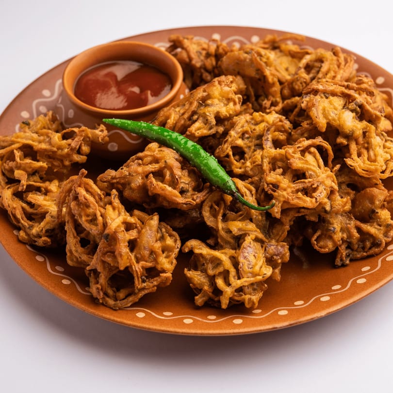 Crispy golden onion pakoras with a side of red chutney and a green chili on a brown plate.