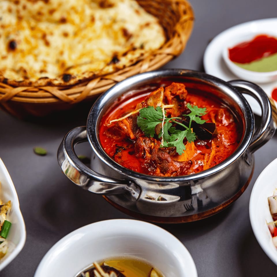 Indian curry in a steel pot with naan bread in a basket and small sauce dishes.
